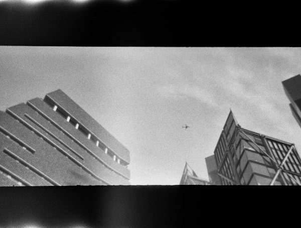 Grainy panoramic black and white photo looking up at several modern city buildings, with an aeroplane in between them