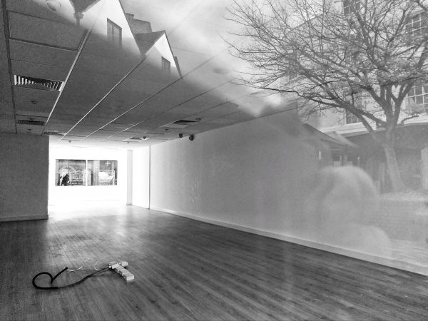 Black and white photo of an empty room with a bright window at the end merging into the reflection of a tree