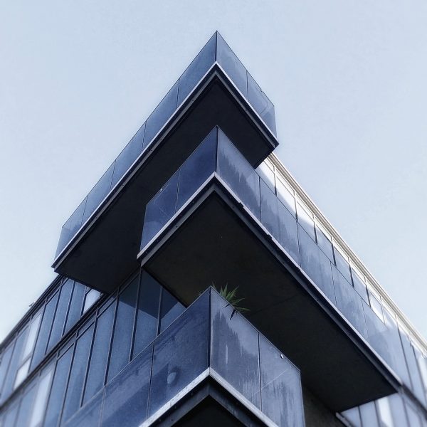 looking up at the corner of a modern-built building with glass balconies overlapping against a clear sky