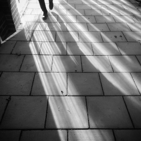 Alternating lines of dark and light reflected across a pavement, with a pair of feet and lower legs just visible at top