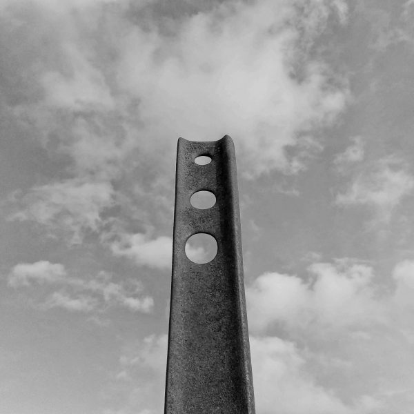 Black and white photo of an abstract, aged, flat metal pillar with three holes through its top end, against a fluffy cloudy sky