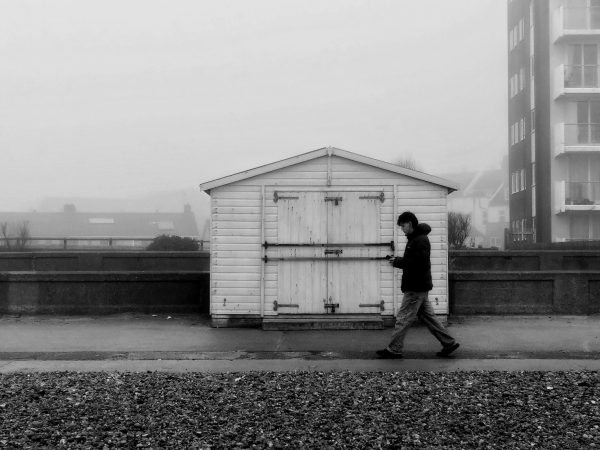 Black and white photo of a man walking in front of a beach hut with the edge of a tower block in the misty background.