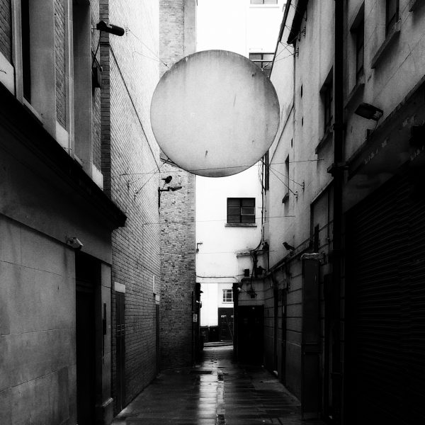 Black and white photo of a narrow London alleyway. Above the middle of the alley hangs a large, white disc ominously taking up the whole width of the lane.