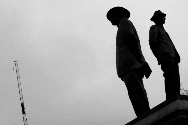 Silhouettes of two statues of men with hats on, facing opposite directions against a blank sky. On the other side, the outline of a metal crane stands tall but forgotten. Do any of them know each other is there?