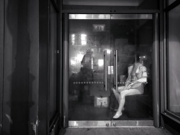 Black and white photo of a shop mannequin body without a head, sitting behind a closed glass door of a shop