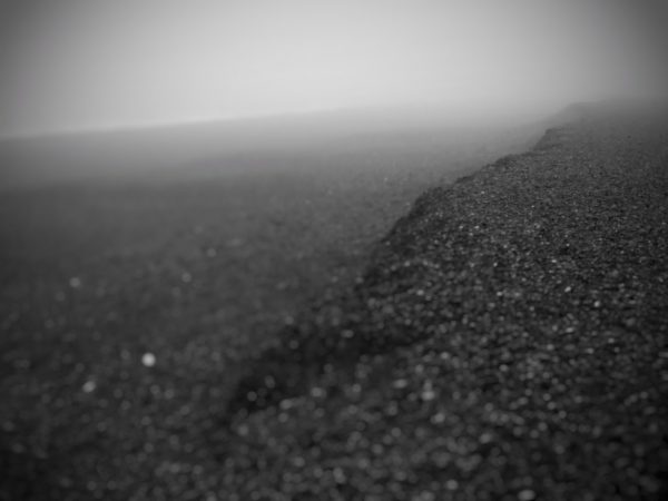 Blurred black and white image showing what appears to be a gravel shoreline receding into mist