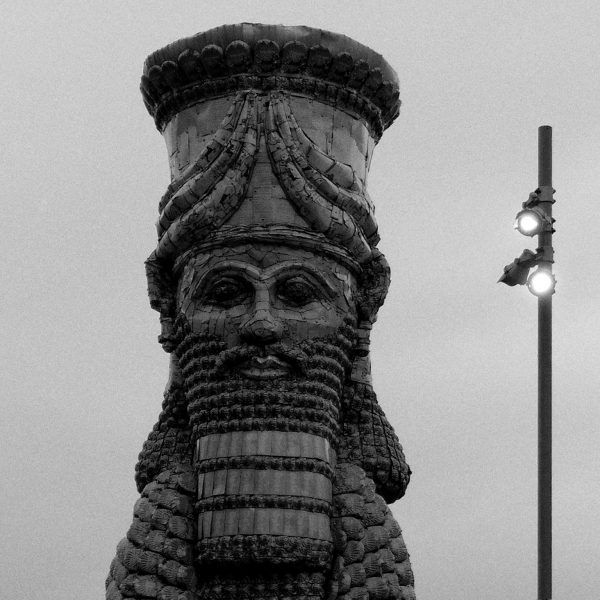 The head of a statue, in the style of the ancient middle east with stylised head gear and beard. A street lamp on a pole shines to the side.