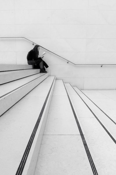 A woman leans against the wall of a staircase reading a book, the parallel lines of the steps converging into a space in front of her very being.