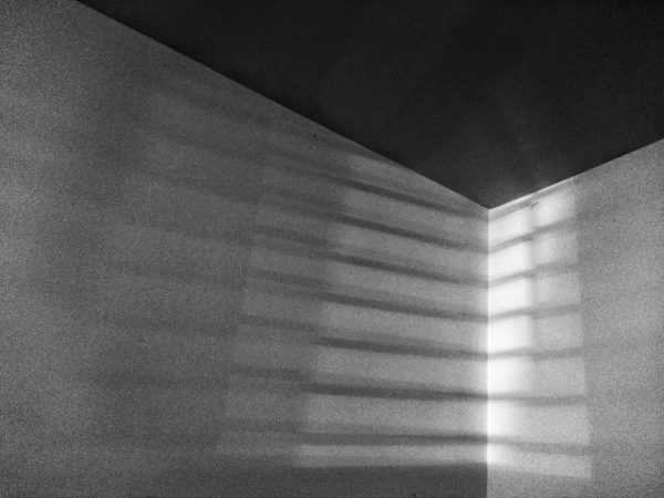 Black and white photo of two walls and a ceiling meeting at a corner with a slatted light pattern cutting across