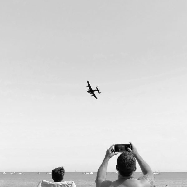 Black and white photo showing the small silhouette of a world war 2 Lancashire bomber in the sky, with the top half of a man in the lower part of the frame holding up a phone screen to take a photo.