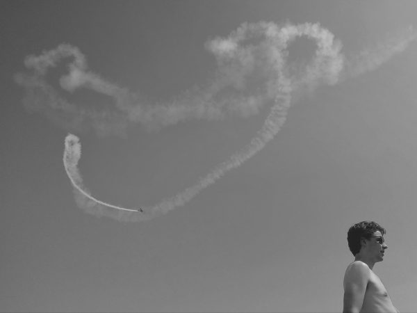 Black and white photo showing an aeroplane and its trail after looping and various acrobatics. A man's head and torso appear in the foreground corner below, facing out of frame.