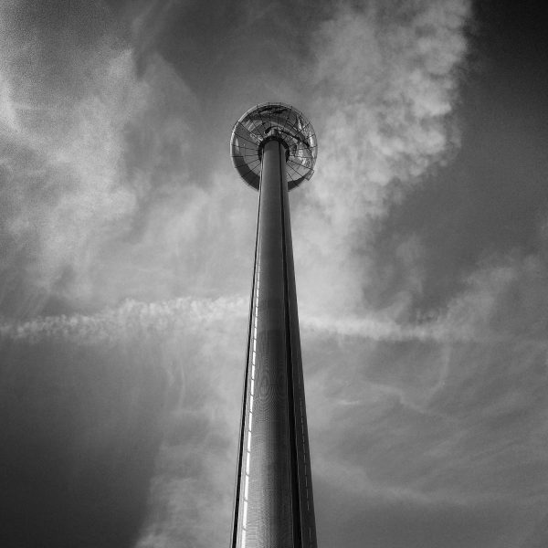 A tall, metal pillar stretches up to a semi-clouded sky, ringed by a glass crown high, high above.