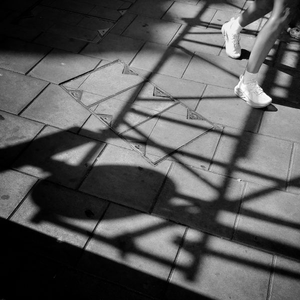 Shadows of scaffolding and signs crisscross a pavement, interrupted only by a pair of legs walking along on white shoes.