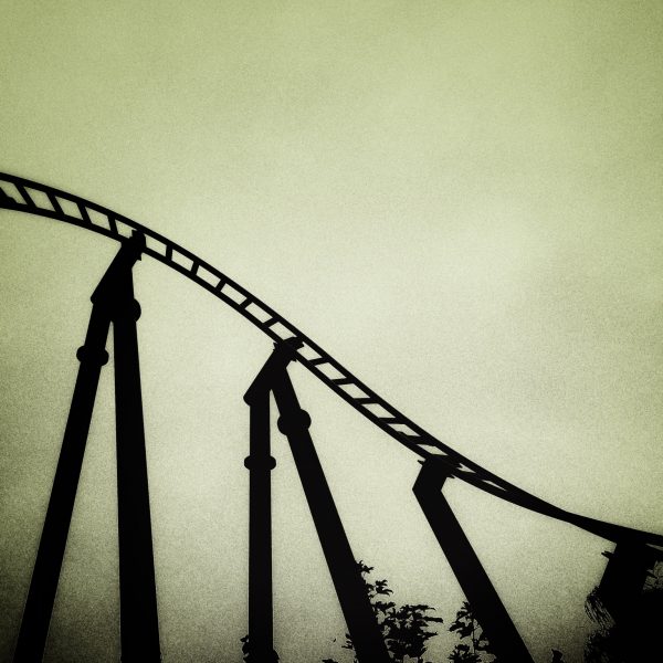 Monochrome photo of the silhouette of an empty rollercoaster rail, propped up by three gantries with plants beginning to encroach upon their stoic sturdiness